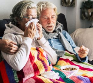 A man and a woman sit on a couch. The woman blows her nose while wrapped in a blanket, and the man's arm is around her shoulders