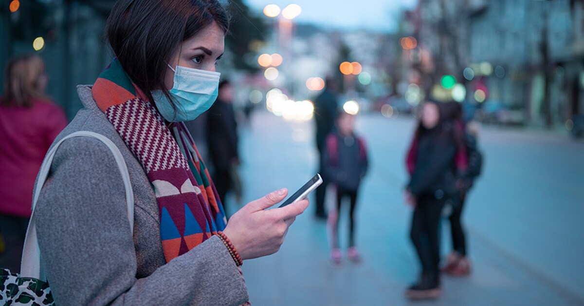 A woman in a mask looks at her phone as she stands on a city street