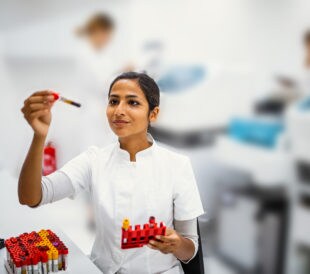 scientist holding up and looking at a sample tube, holding rack of sample tubes.