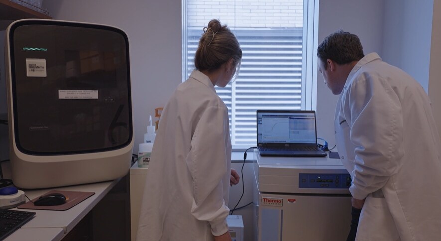 Researchers in lab coats reviewing qPCR data on a Thermo Fisher instrument in a molecular biology lab.