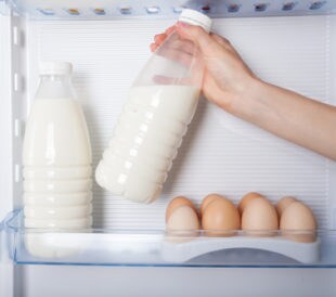 A hand reaches into a fridge to grab one of two milk bottles that are next to a dozen eggs. Image: takoburito/Shutterstock.com