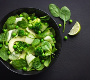 A salad composed of peas, spinach, broccoli and melon sits in a black bowl on a black table.