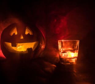 Halloween pumpkin with carved face and glass of whiskey with ice on a dark toned foggy background with zombies. Decorated. Selective focus