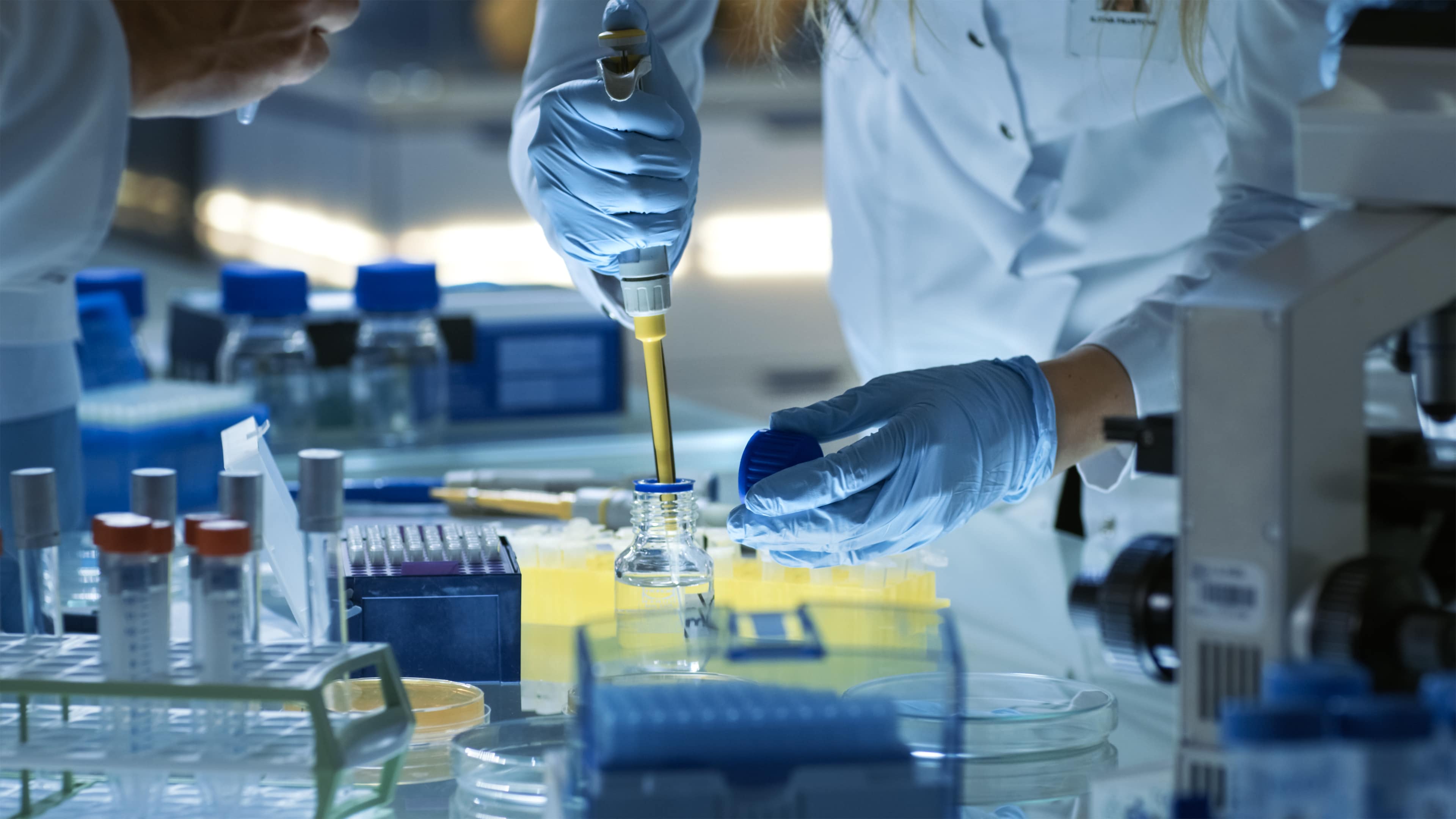 A lab tech's hand holds a pipette over a bottle in a lab