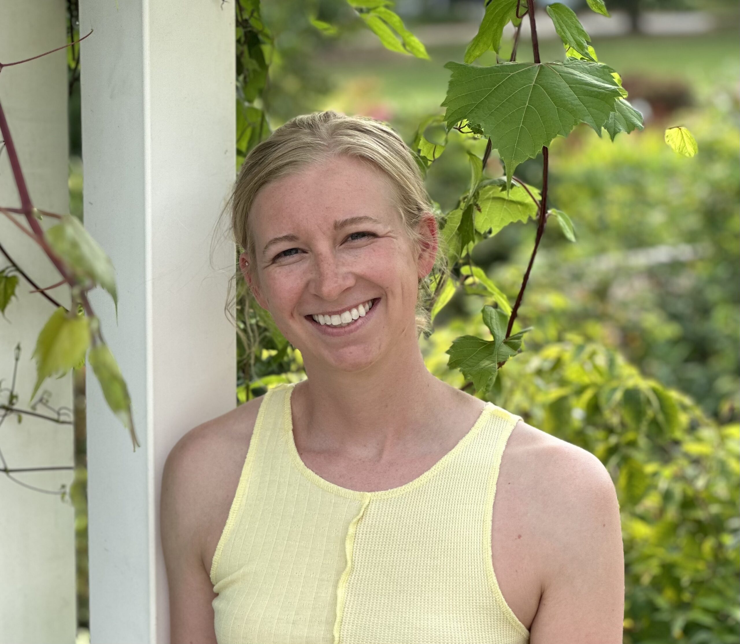 Portrait of a smiling plant pathology researcher standing outdoors beside grapevine leaves.